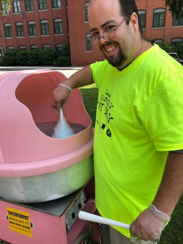 A smiling white male with black, short hair, mustache, and beard. He is wearing a neon green shirt with black unreadable text. He is spinning blue cotton candy in a pink machine.